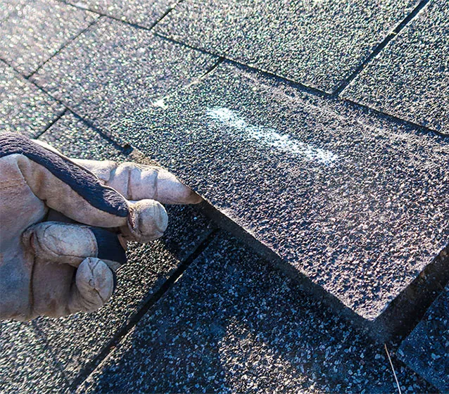 Gloved hand lifting a damaged asphalt shingle